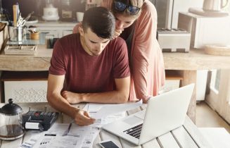 Young family managing budget, reviewing their bank accounts using generic laptop pc and calculator in kitchen. Husband and wife doing paperwork together, paying taxes online on notebook computer